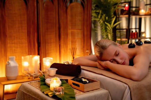 Hot stone massage at spa salon in luxury resort with warm candle light, blissful woman customer enjoying spa basalt stone massage glide over body with soothing warmth. Quiescent