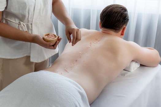 Blissful man customer having exfoliation treatment in luxury spa salon with warmth candle light ambient. Salt scrub beauty treatment in health spa body scrub. Quiescent