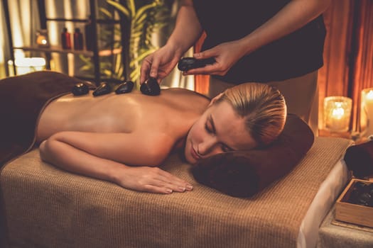 Hot stone massage at spa salon in luxury resort with warm candle light, blissful woman customer enjoying spa basalt stone massage glide over body with soothing warmth. Quiescent