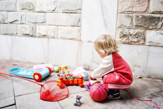 Little girl is playing with different colorful toys while squatting against a stone wall. High quality photo