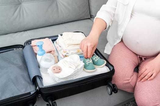 A pregnant girl packs her things and cute little shoes into a suitcase.