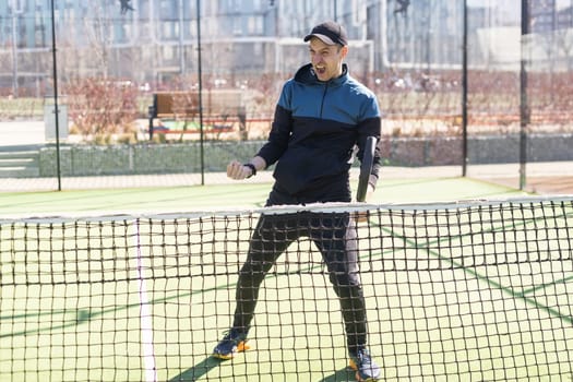 A padel player jump to the ball, good looking for posts and poster. Man with black racket playing a match in the open behind the net court outdoors. Professional sport concept with space for text. High quality photo