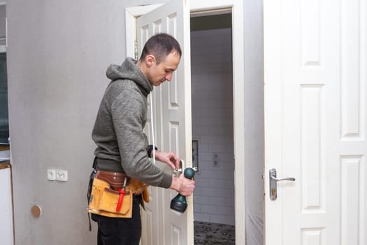 Maintenance Concept. Focused serious young locksmith with tattoo on hand wearing blue uniform standing on one knee in doorway installing doorknob handle, repairing wooden front door entrance. High quality photo