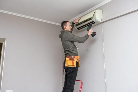 Professional electrician man with screwdriver maintaining, cleaning modern air conditioner indoors. Young technician standing on a ladder in the room repairing or installing air conditioner. High quality photo
