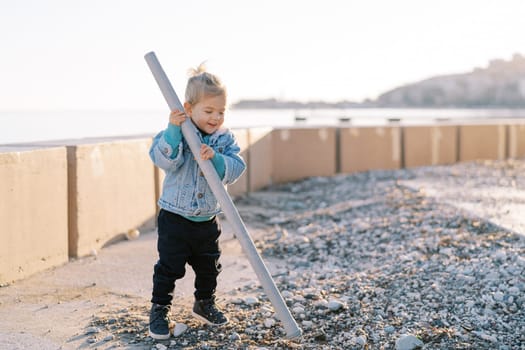 Little laughing girl picking pebbles with a plastic pipe while standing near the fence on the seashore. High quality photo