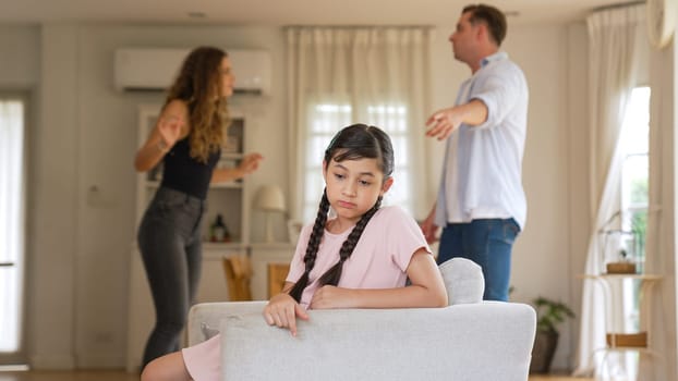 Annoyed and unhappy young girl sitting on sofa trapped in middle of tension by her parent argument in living room. Unhealthy domestic lifestyle and traumatic childhood develop to depression Synchronos