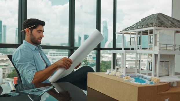 Smiling engineer looking project plan while design house model at office. Smart caucasian sitting relaxing while reading project plan. Creative design, civil engineering. Skyscraper. Tracery