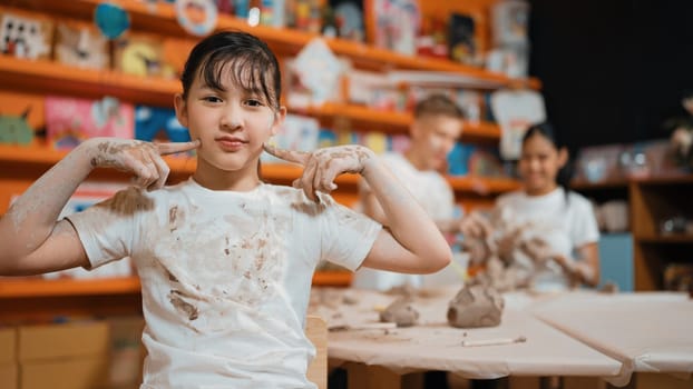 Asian girl pose at camera while diverse children modeling clay behind. Happy cute student wearing dirty shirt while looking at camera at workshop in art lesson. Blurring background. Edification.