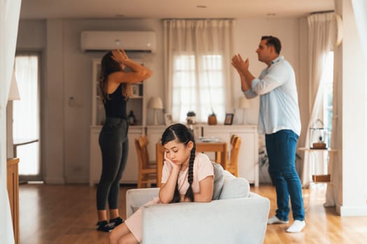 Annoyed and unhappy young girl sitting on sofa trapped in middle of tension by her parent argument in living room. Unhealthy domestic lifestyle and traumatic childhood develop to depression.Synchronos