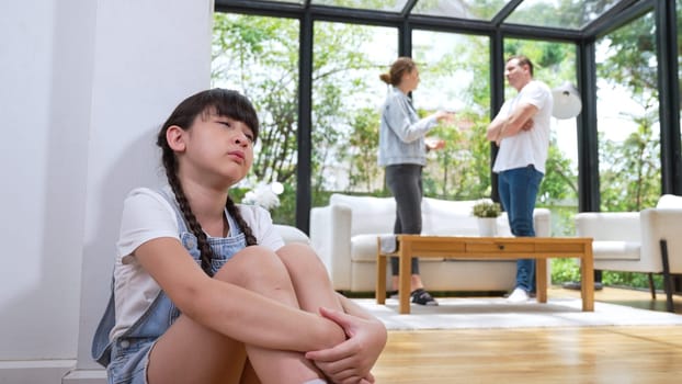 Stressed and unhappy young girl huddle in corner, cover her ears blocking sound of her parent arguing in background. Domestic violence at home and traumatic childhood develop to depression. Synchronos