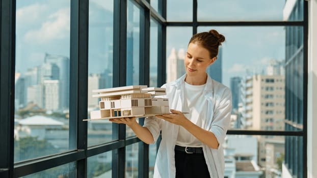 Young smart caucasian beautiful architect engineer raised house model while inspect carefully surrounded by city skyscraper view. Civil engineering, architectural studio. project planing. Tracery