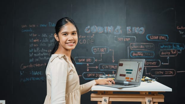 Caucasian girl working on laptop while smiling to camera at blackboard with engineering code and prompt written. Skilled pretty academic student learning programing or coding system. Edification.