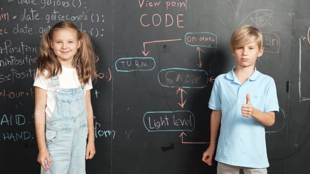 Caucasian girl standing at blackboard with engineering prompt or coding, programing system written in STEM technology classroom. Cute student looking at camera while boy showing thumb. Erudition.