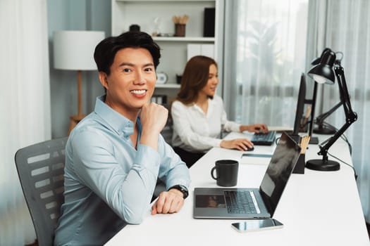 Profile smiling of smart Asian businessman looking camera to pose at modern office. Blurry background woman colleague working on pc to check email marketing plan analysis report at desk. Infobahn.