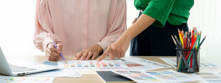 Cropped image of two female young beautiful graphic designer choose the color from color palate by using laptop on table with work tools and equipments scatter around at modern office. Variegated.