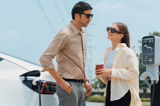 Young couple with coffee recharge EV car battery at charging station connected to electrical industrial power grid tower. Couple with shopping and travel using eco electric car lifestyle. Expedient