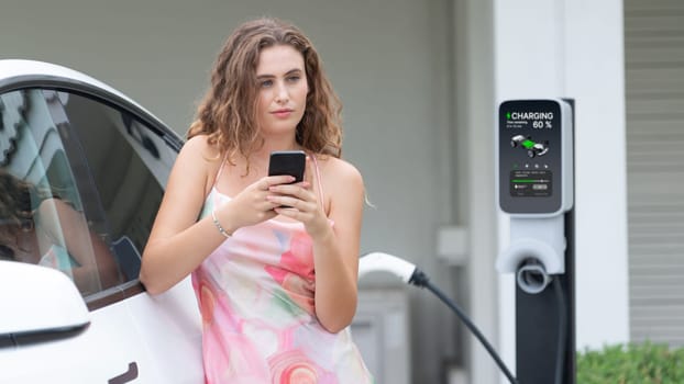 Modern eco-friendly woman recharging electric vehicle from home EV charging station. Innovative EV technology utilization for tracking energy usage to optimize battery charging at home. Synchronos