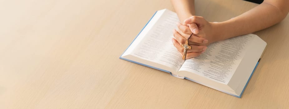 Cropped image of praying male hand holding cross on holy bible book at wooden table. Top view. Concept of hope, religion, faith, christianity and god blessing. Warm and brown background Burgeoning.