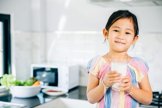 Cute Asian little girl in kitchen holds milk smiling joyfully. Portrait of daughter enjoying drink at home. Happy preschooler savoring calcium-rich liquid radiating happiness give me.
