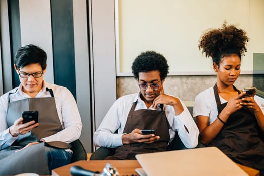 A diverse group of baristas sitting together in a cafe looking at smartphones enjoying relaxation. Team of professionals in uniform connecting through phones. Teamwork and connection evident. No order