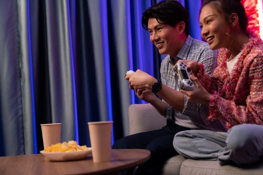 Sitting sofa of happy smiling couple playing video game control by joystick fronted snack and drinks competing challenge level with funny at modern neon blub light comfy living room at home. Infobahn.