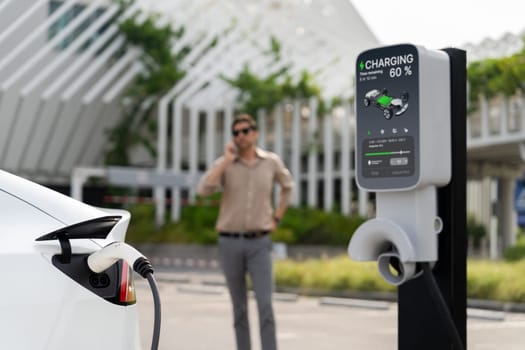 Young man recharge EV electric vehicle at green city commercial mall parking lot while talking on phone. Sustainable urban lifestyle for eco friendly EV car with battery charging station. Expedient