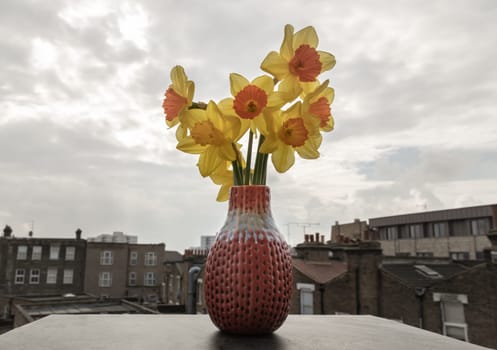 Bunch of daffodils in a red ceramic vase on the terrace with sky background. Arrangement of Yellow Spring Flowers Daffodils, Amazing view background with Yellow flowers, Space for text, Selective focus.