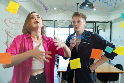 Couple of businesspeople discussing and brainstorming about solving marketing problem in front of glass board at business meeting by using mind map and sticky notes. Working together. Immaculate.