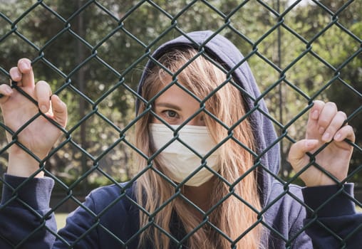 Close-up shot of young woman wearing Protective Face Mask stand behind fence border. COVID-19 coronavirus infection pandemic disease virus