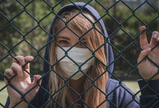 Close-up shot of young woman wearing Protective Face Mask stand behind fence border. COVID-19 coronavirus infection pandemic disease virus