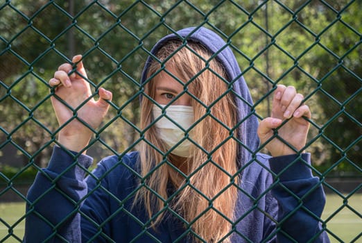 Close-up shot of young woman wearing Protective Face Mask stand behind fence border. COVID-19 coronavirus infection pandemic disease virus