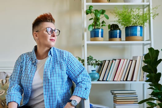 Portrait of middle-aged woman with red hair wearing glasses in casual clothes sitting on couch at home looking out window, copy space