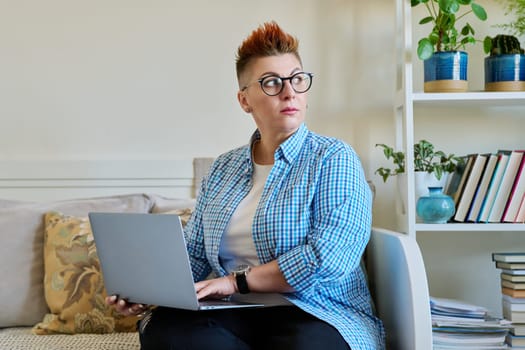 Middle aged woman sitting on couch at home using laptop. Serious female with red haircut in casual clothes working on computer, relaxing, communicating