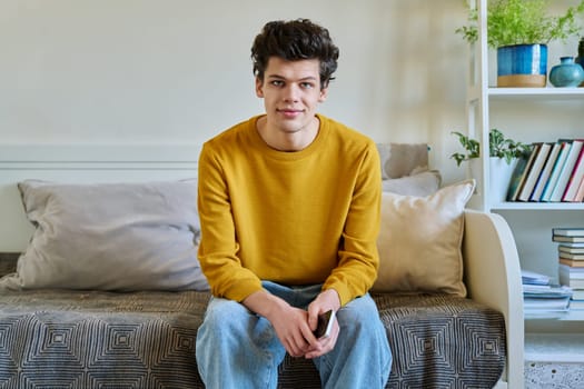 Portrait of handsome smiling guy looking at camera, sitting on couch at home. Young man in yellow with curly hair, university college student. Lifestyle, youth 19-20 years old concept