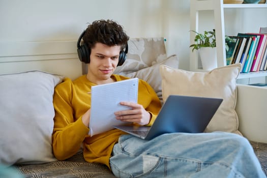 Young guy college student in headphones using laptop making notes, sitting on couch at home. Webinar listening audio online conference chat lesson e-learning education remote classes internet meetings