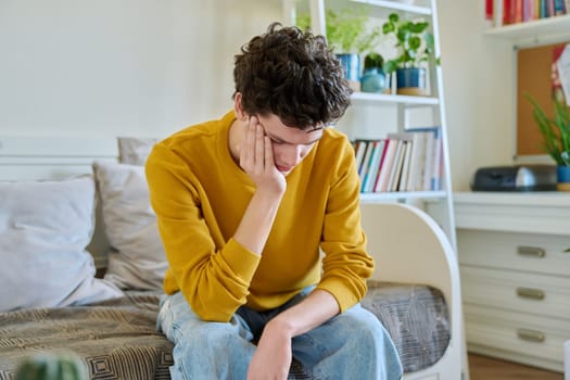 Sad upset tired young man sitting on couch at home, touching his head with hands. Health problems, headaches, troubles, difficulties in study, family relationships, mental health, stress, depression