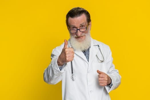 Like. Senior elderly doctor cardiologist man raises thumbs up agrees, gives positive reply good idea feedback celebrating success victory. Apothecary pharmacy grandfather isolated on yellow background