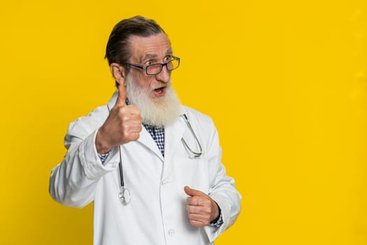 Like. Senior mature doctor cardiologist man raises thumbs up agrees, gives positive reply good idea feedback celebrating success victory. Apothecary pharmacy grandfather isolated on yellow background