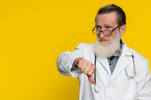 Dislike. Upset unhappy senior doctor cardiologist man showing thumbs down sign gesture disapproval dissatisfied negative feedback. Apothecary pharmacy grandfather on yellow background. Copy-space