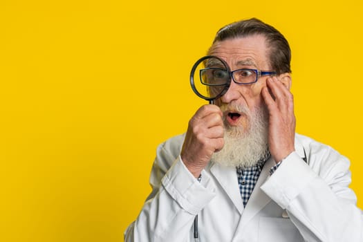Investigator researcher scientist senior doctor cardiologist man holding magnifying glass near face looking, searching, analyzing. Apothecary pharmacy grandfather on yellow background. Copy-space