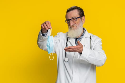 Upset elderly doctor cardiologist man removing taking off medical protective mask. Flu treatment cure. Ending of pandemic quarantine. Apothecary pharmacy old grandfather isolated on yellow background