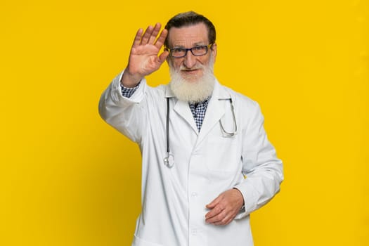Senior smiling doctor cardiologist man waving hands gesturing hello greeting or goodbye welcoming with invitation hospitable positive expression. Apothecary pharmacy grandfather on yellow background