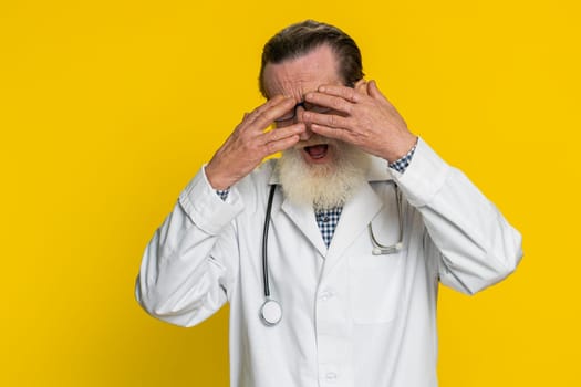 Don't want to look at this awful. Afraid shocked senior elderly doctor cardiologist man close eyes with hands confused shy scared reject to watch. Apothecary pharmacy grandfather on yellow background