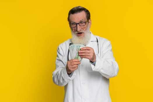 Rich happy senior doctor cardiologist man waving money dollar cash banknotes bills like a fan, success business career, big income wealth. Apothecary pharmacy grandfather isolated on yellow background