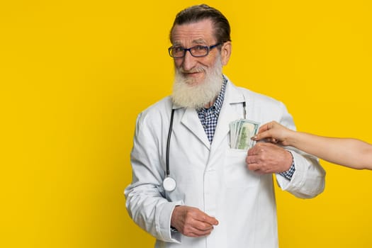 Senior speaker doctor cardiologist man receiving bribe from patient taking money from client hiding cash into pocket. Illegal corruption in medicine. Sneaky apothecary grandfather on yellow background