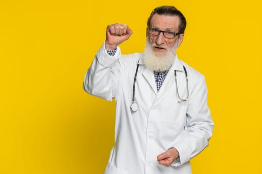 Knock-knock who is there. Excited senior doctor cardiologist man knocking door gesture asking who is at home being clueless uncertain. Hello, welcome. Apothecary old grandfather on yellow background
