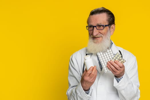 Senior doctor cardiologist man shows lot of tablet pills in blisters recommends drugs vitamin antibiotic cure treatment. Health care. Apothecary pharmacist grandfather isolated on yellow background