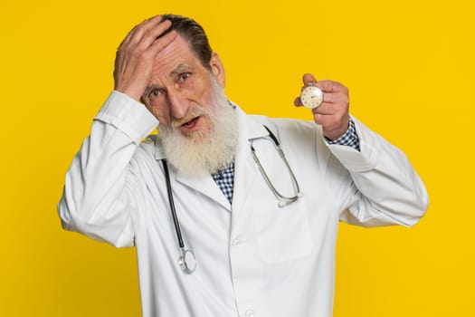 Mature doctor cardiologist man with anxiety checking time on pocket watch, running late to work being in delay deadline. Pharmacy old grandfather worrying to be punctual isolated on yellow background