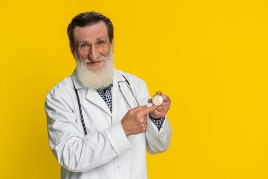 It is your time to heal. Senior elderly doctor cardiologist man showing time on pocket watch clock ok thumb up approve pointing finger at camera. Apothecary pharmacy grandfather on yellow background