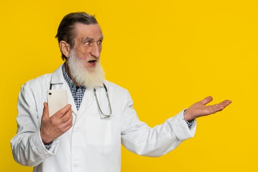 Mature doctor cardiologist man using smartphone showing pointing empty place, advertising application area, positive feedback, copy-space. Apothecary pharmacy grandfather isolated on yellow background